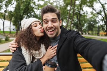 Portrait of brunette couple man and woman 20s taking selfie photo, while sitting on bench in green park