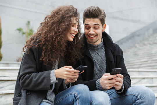 Image of brunette couple man and woman 20s in warm clothes, using smartphones together while sitting on stairs outdoor
