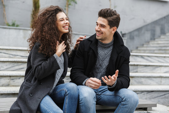 Image of joyful couple man and woman 20s in warm clothes, laughing while sitting on stairs outdoor