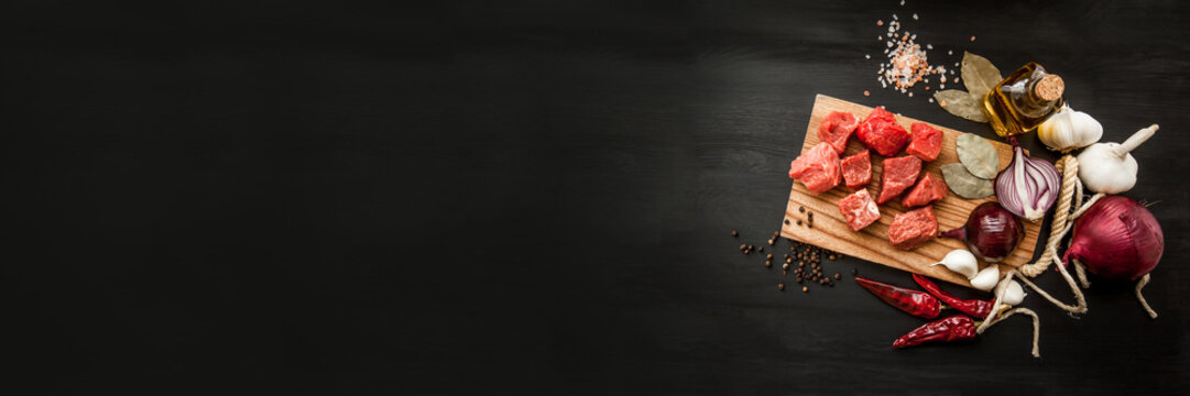 Uncooked fresh diced beef meat with herbs and oil on an old rustic wooden kitchen board over black background. Top view with free space for text. Banner