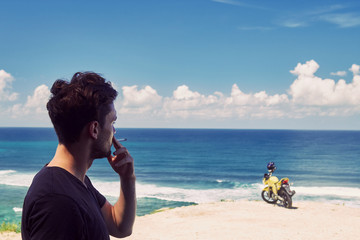 Young handsome man smoking cigar while standing near motorcycle on the tropical beach.