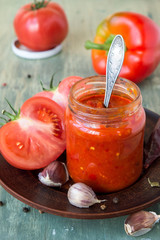 A glass jar with freshly prepared Georgian adjika sauce, a red ripe tomatoes, pepper and garlic on an old wooden table. Selective focus.