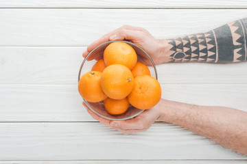 cropped view of tattooed man holding glass bowl with ripe tangerines on wooden surface