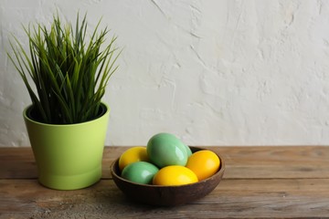 Colorful easter eggs in a brown bowl, grass in a green flowerpot. Wooden table, light background. Copy space, horizontal orientation