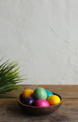 Colorful eggs in a brown bowl, grass. Wooden table, light background. Copy space, vertical orientation