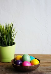 Colorful easter eggs in a brown bowl, grass in a green flowerpot. Wooden table, light background. Copy space, vertical orientation