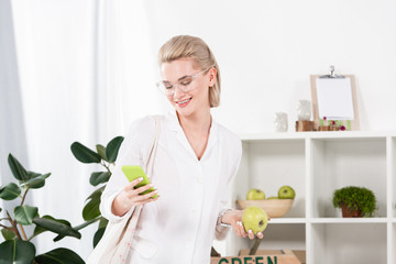 cheerful businesswoman in glasses holding green apple and using smartphone