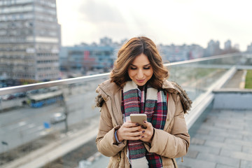 Portrait of beautiful brunette in jacket and with scarf standing on rooftop and using smart phone.