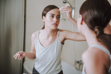 Serious nice woman applying face gel in front of the mirror