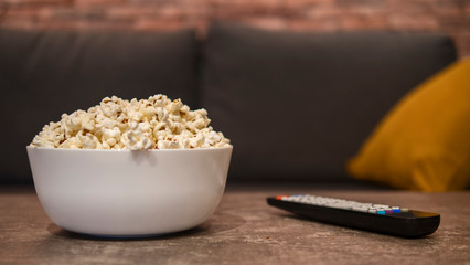 White bowl of white salty popcorn and remote control on a wooden table in front of a couch. Front view, blurry background