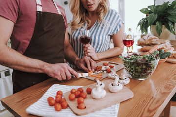 Blond lady with drink watching how her husband cooking dinner