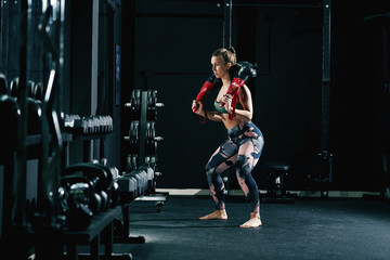 Fototapeta na wymiar Muscular strong caucasian female bodybuilder in sportswear and with pigtail doing exercises with bulgarian training bag barefoot.