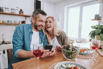 Cheerful couple using cellphones and having dinner at home