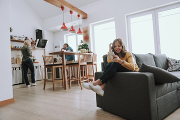 Cute little girl sitting on comfortable gray couch and using cellphone at home