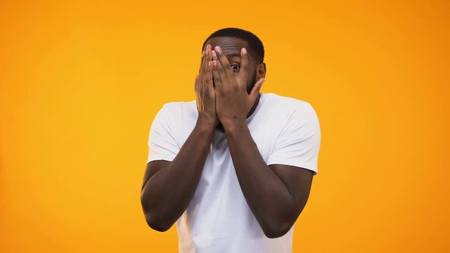 Surprised afro american guy peeping through fingers against yellow background