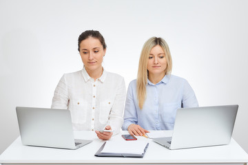 Fototapeta na wymiar Beautiful brunette female manager with a smile and her blonde caucasian colleague works with laptops sits by the desk in the office with the white background.