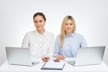 Beautiful brunette female manager with a smile and her blonde caucasian colleague watches straight sits by the desk in the office with the white background.