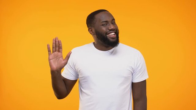 Affable young black man waving hand neighborly, isolated on yellow background