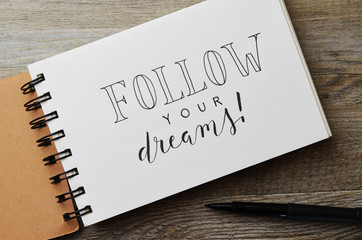 FOLLOW YOUR DREAMS! hand-lettered in notebook on wooden desk with pen