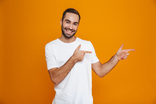 Image of smiling guy 30s in t-shirt pointing fingers aside while standing, isolated over yellow background