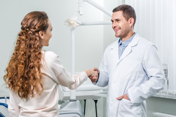 cheerful dentist standing with hand in pocket and shaking hands with female patient