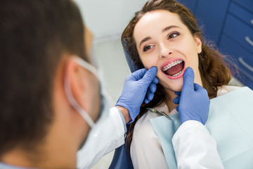 selective focus of woman in braces opening mouth during examination of teeth near dentist - obrazy, fototapety, plakaty