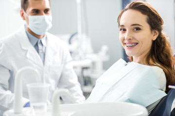 selective focus of woman in braces smiling with dentist standing in mask on background