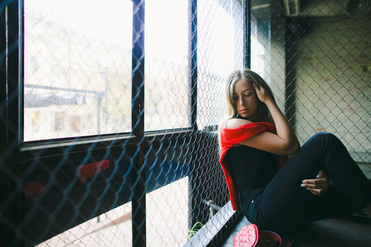 Streetstyle female portrait. Woman dressed in red shirt and sport pands sits in the dark room