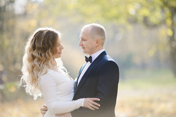 Bride and groom hug each other tender standing in the rays of sun in the autumn park covered with fallen leaves