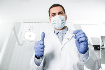 dentist in latex gloves and mask holding dental instrument and showing thumb up
