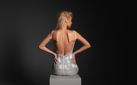 High fashion portrait of young elegant woman. Young lady in amazing silver glitter sequins luxury dress sitting on  armchair Studio shot. Back view 