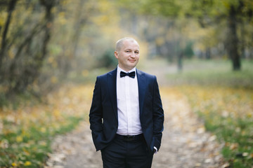 Handsome groom in classy black suit poses in autumn park covered with fallen leaves
