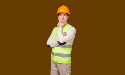 The thoughtful man in a construction helmet and a vest, looks in the camera, the crossed hands at the head