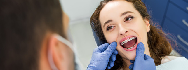 selective focus of cheerful woman in braces opening mouth during examination of teeth near dentist