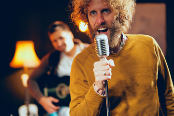 Close up of young Caucasian man with curly hair holding microphone and singing. Home studio interior.