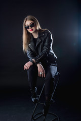 young girl in a black leather jacket and sunglasses on a dark background in the studio sitting