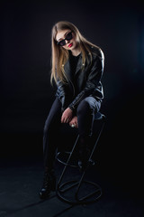 young girl in a black leather jacket and sunglasses on a dark background in the studio sitting