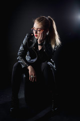 young girl in a black leather jacket and glasses on a dark background in the studio sitting