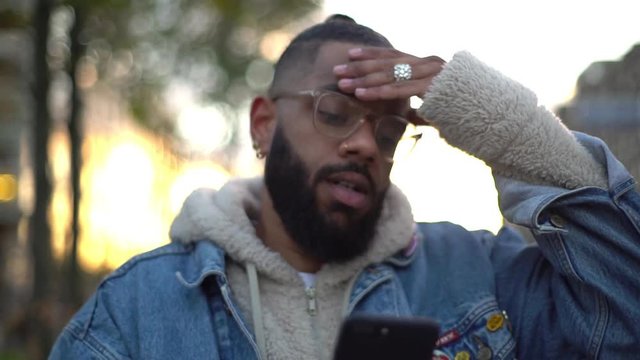 Slow-mo attractive black guy wearing glasses, hairbun, looking puzzled, confused smartphone shaking head disagree, smirking tensed disappointed, perplexed find address, searching hostel foreign city