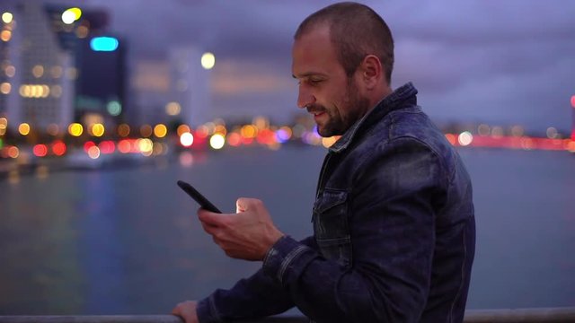 Slow-mo happy handome caucasian bearded guy standing bridge return home evening stop near river send girlfriend photo city view shot looking smartphone display scrolling pics smiling happily
