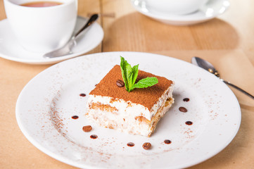 Delicious Tiramisu cake with coffee beans and fresh mint on a plate on a light wooden background. Selective focus, copy space.