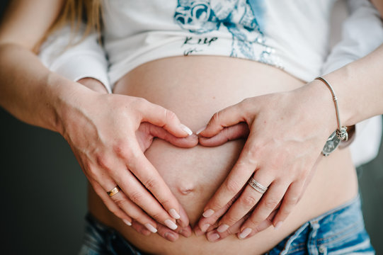 Cropped image of husband holding belly of his pregnant wife making symbol heart hands. Pregnant woman and loving handsome man hugging tummy at home. Loving Couple. Parenthood concept. Baby Shower.