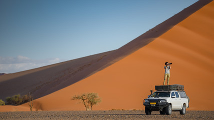 Fototapeta na wymiar Young Asian man traveler and photographer standing on camper car near orange sand dune. Desert road trip travel concept