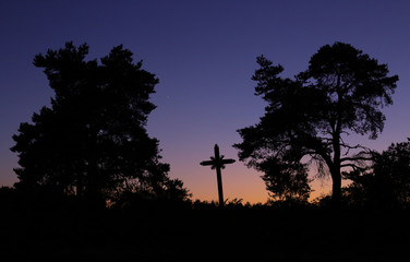 Black silhouette of old wooden cross and pine trees in front of purple evening sky after sunset....