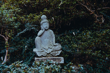 A weathered stone statue of a Buddha or Buddhist monk sitting and observing world, meditates under green trees in japanese garden. Relax and mind calm concept. Exterior,outdoor decor. Copy space.