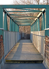a perspective view along an green rusting metal pedestrian footbridge with rails and girders casting shadows in afternoon sunlight with trees in the background