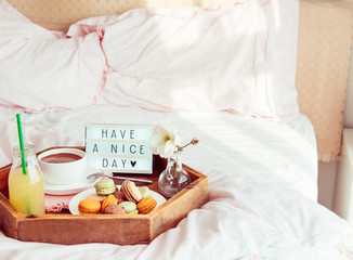 Fototapeta na wymiar Breakfast in bed with Have a nice day text on lighted box. Cup of coffee, juice, macaroons, flower in vase on wooden tray. Good morning mood. Hospitality, care, service concept. Copy space.