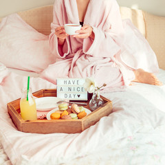 Fototapeta na wymiar Good morning mood. No face woman in bathrobe sitting on the bed with coffee and has breakfast in bed with Have a nice day text on lighted box. Hospitality, care, service concept. Square. Copy space.
