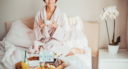 Fototapeta na wymiar Good morning mood. No face woman in bathrobe sitting on bed with coffee and has breakfast in bed with Have a nice day text on lighted box. Hospitality, care, service concept. Wide banner. Copy space.