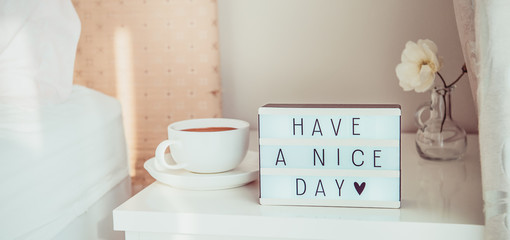 Close up Have a nice day text message on lighted box, cup of coffee and white flower on the bedside table in sun light. Good morning mood. Hospitality, care, service concept. Wide banner. Copy space.